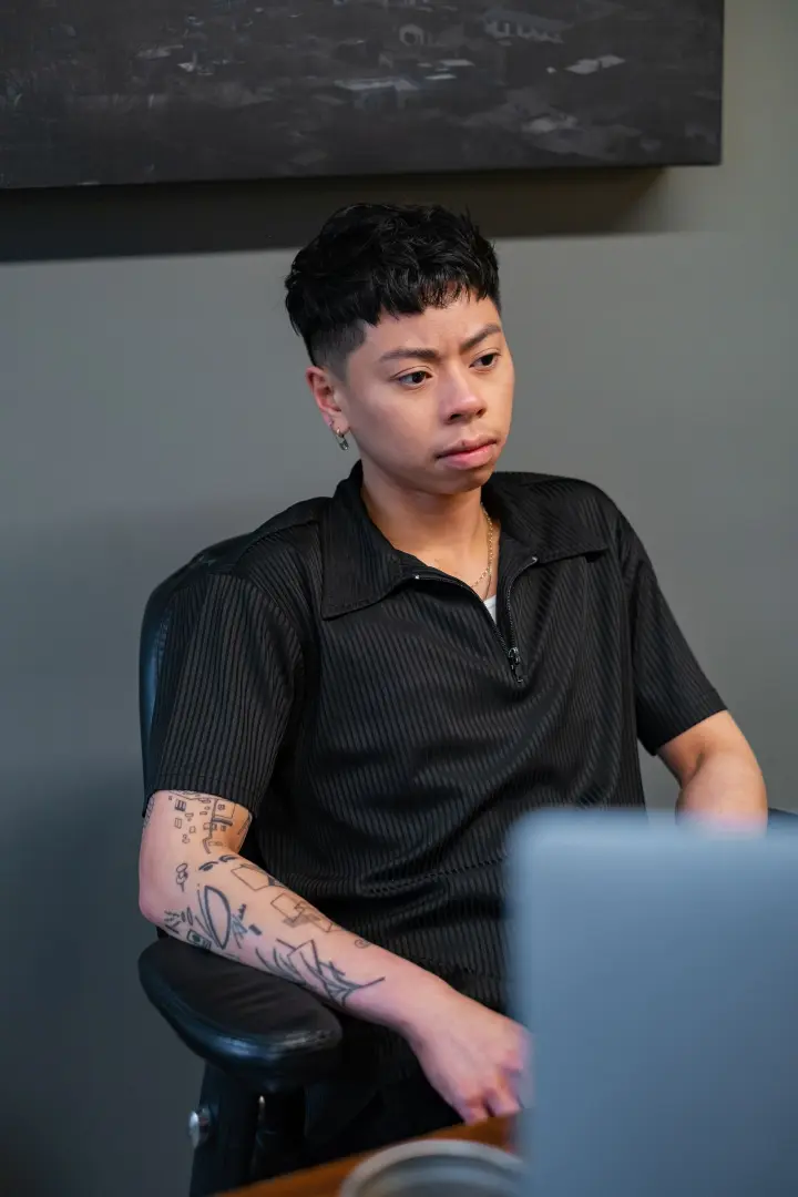A non-binary person sitting behind a desk, looking frustrated or unsure.