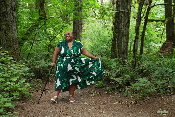 In a lush green forest, a Black non-binary person smiles while twirling the skirt of their flowing leaf print dress and dancing with their cane in hand. Their head is shaved and they wear glasses, gold earrings, and gold sandals.