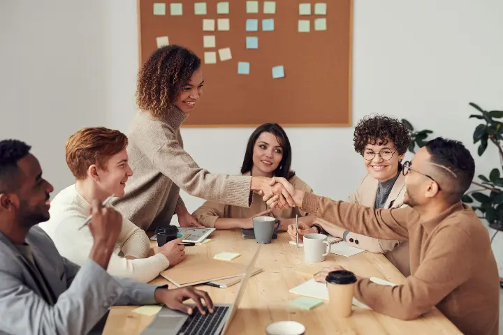 A group of diverse folks in a meeting, focusing on two colleagues shaking hands. They look happy and comfortable with each other's presence.