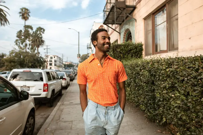 A smiling person wearing an orange shirt, walking leisurely on a sidewalk.