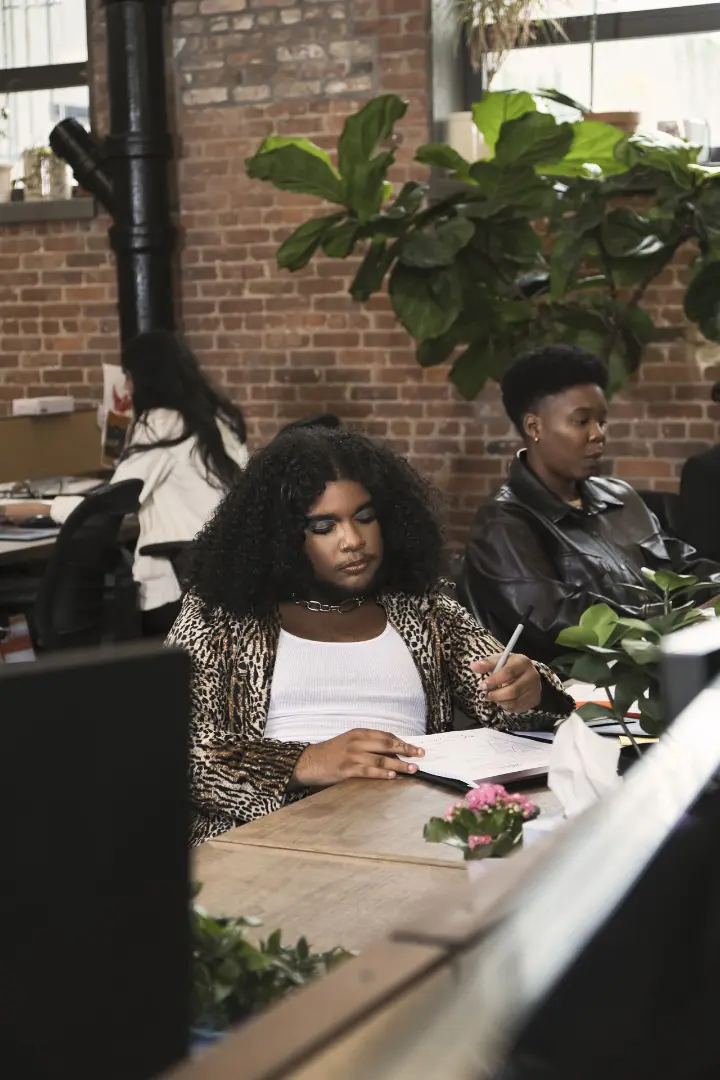 A group of people of varying genders work in an office, with a focus on a non-binary person writing on a notebook.