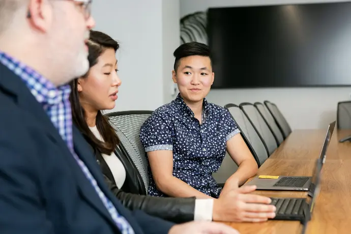 A queer leader is in focus, smiling towards a team member who's presenting to the group.