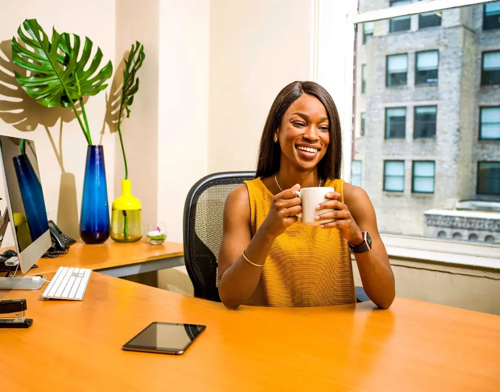 A Latinx person sitting on their desk, smiling widely while holding a mug.