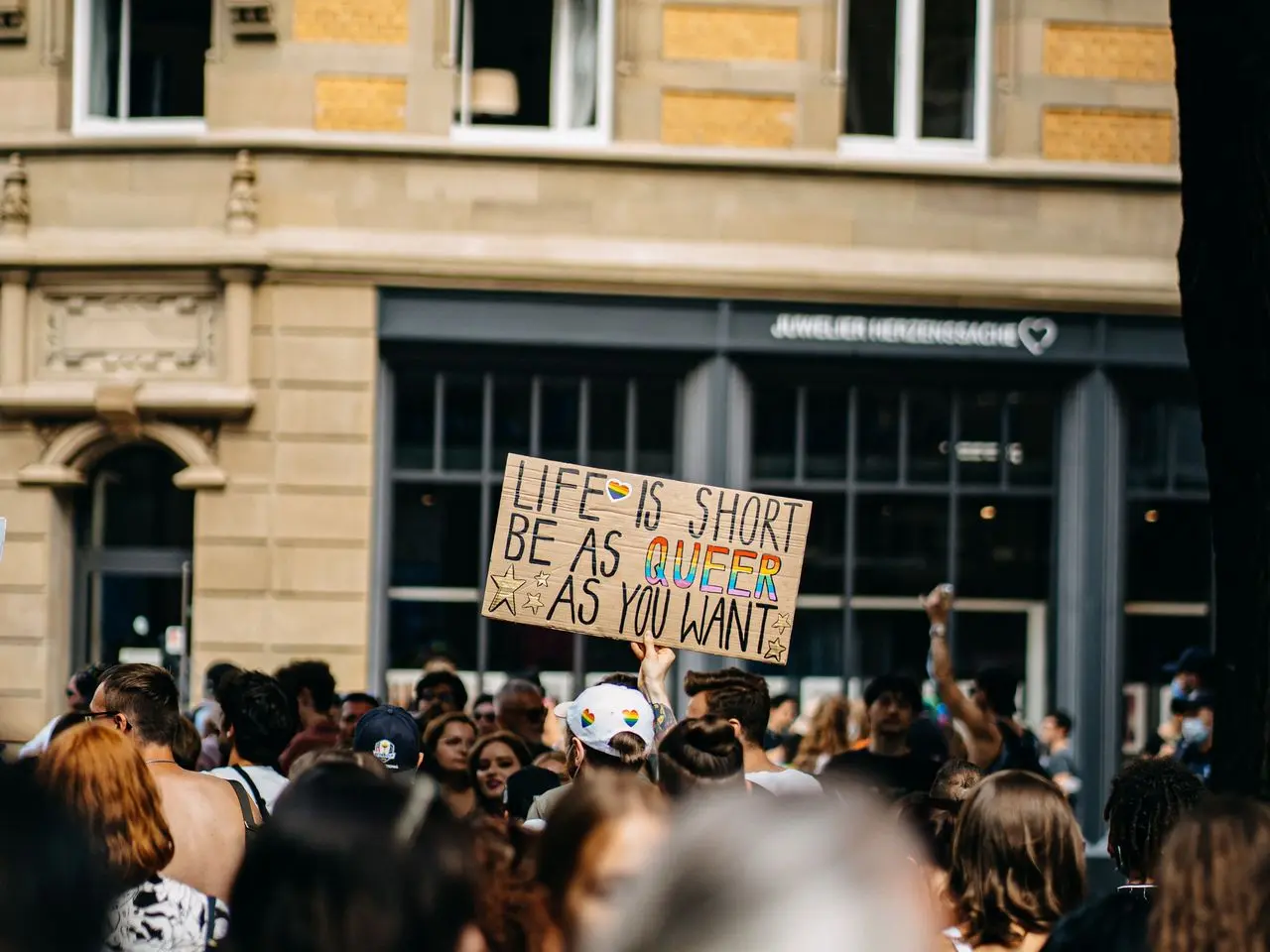 Daytime photo of a person in a Pride parade holding a signage that reads "Life is short, be as queer as you want."