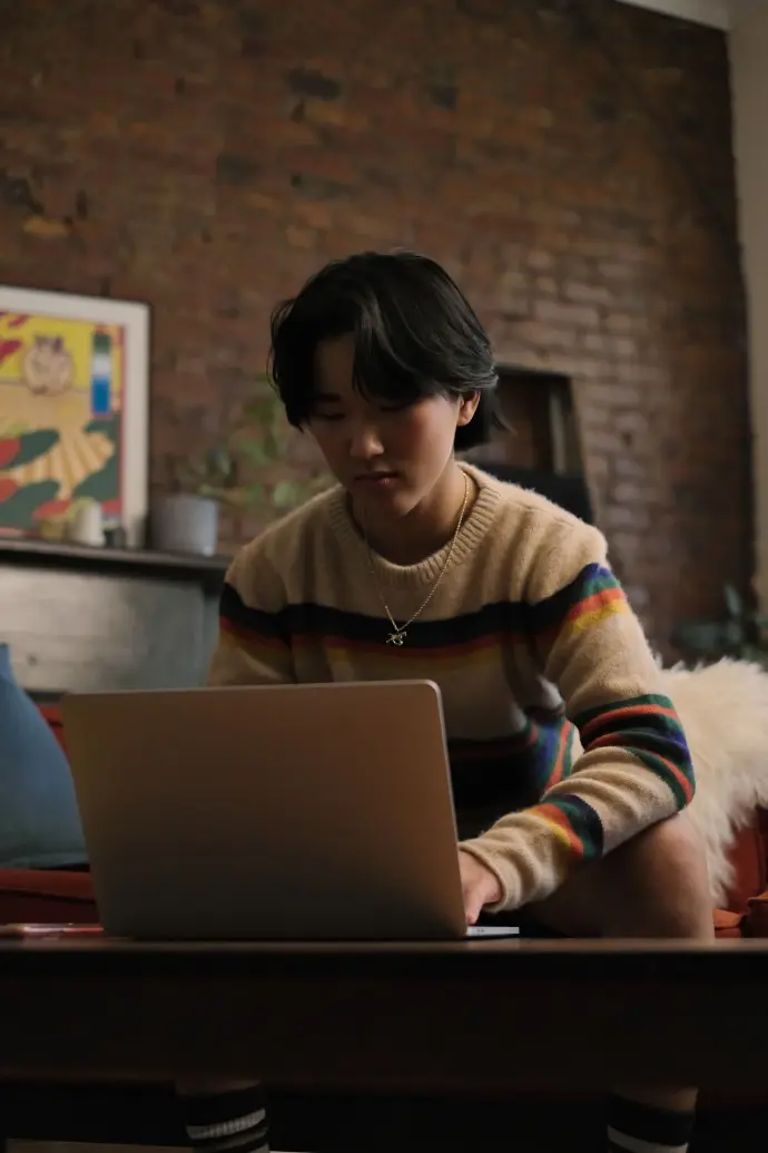 A nonbinary person looking pensive while seated on a soft sofa and typing on a laptop. They are wearing a multicolored sweater and a gold chain necklace with a horse pendant. A brick wall, together with a pop art painting and a plant over a wooden shelf, serve as backdrop.