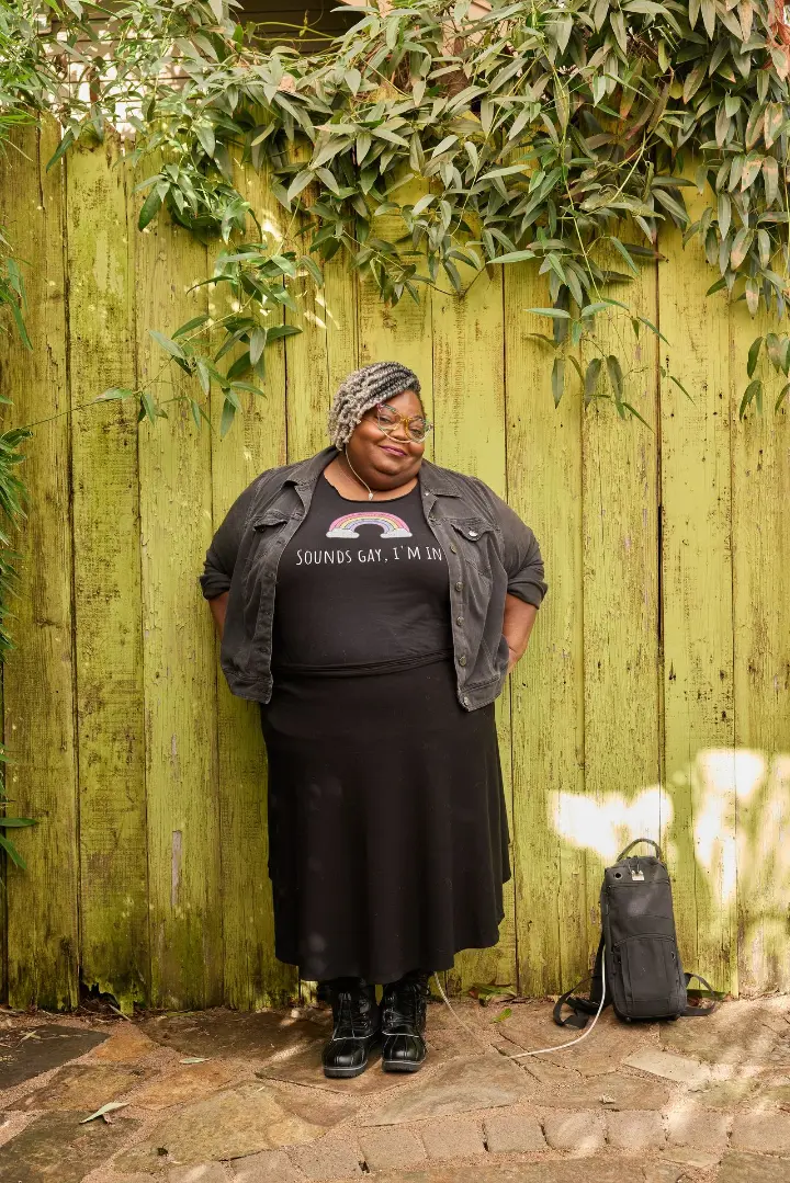 A Black woman in cat eye glasses and a nasal cannula smiles at the camera while standing next to her portable oxygen backpack. She has ombré gray twists and is in an all-black outfit, with a denim jacket, long skirt, boots, and a rainbow shirt declaring, “Sounds gay, I’m in.”